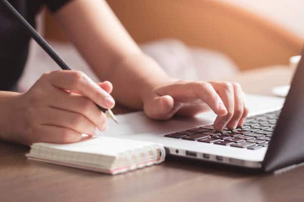 Gros plan sur les mains d'une femme qui tape sur un clavier d'ordinateur et écrit sur un petit bloc-note